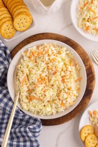 Classic coleslaw in a white serving bowl on a wood board with small plates, crackers, and a blue gingham napkin.
