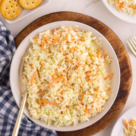 Classic coleslaw in a white serving bowl on a wood board with small plates, crackers, and a blue gingham napkin.