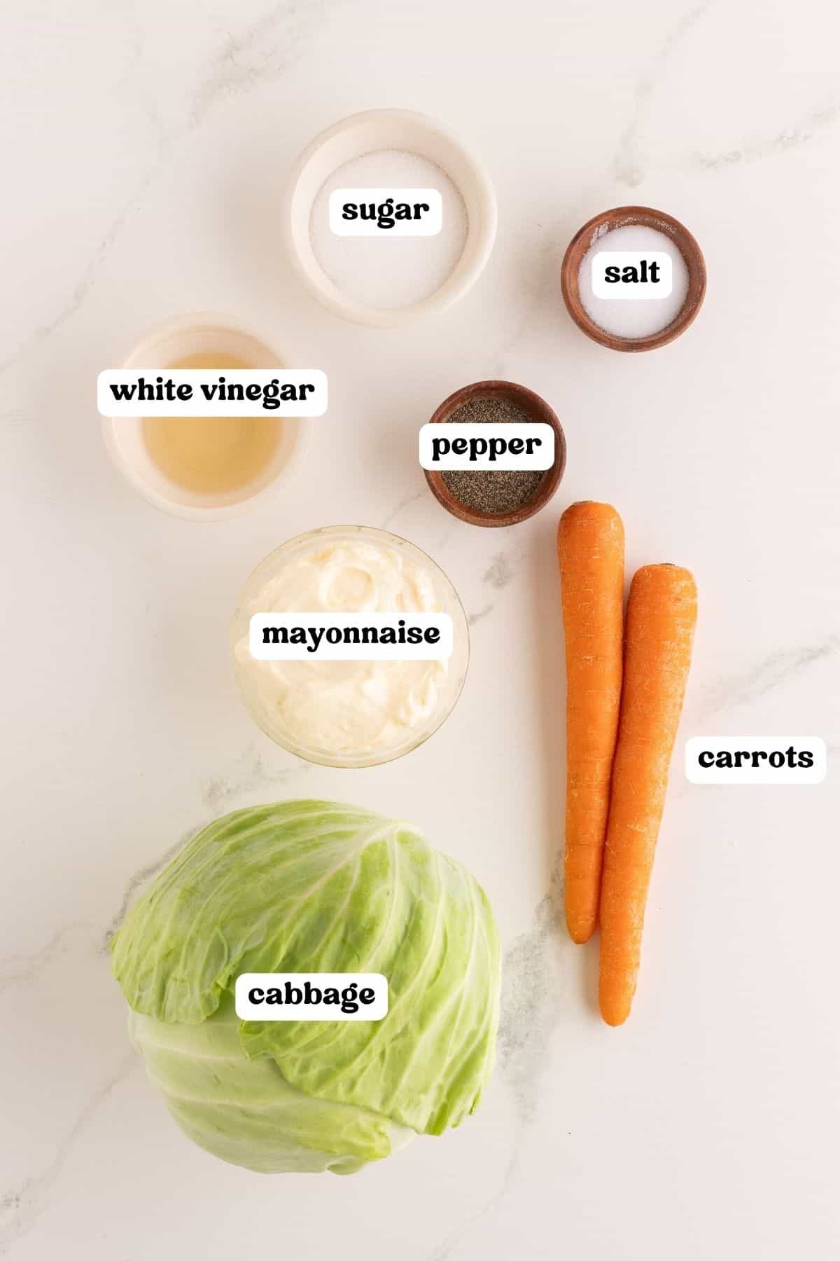 Ingredients on a marble counter: a head of green cabbage, two carrots, white vinegar, mayonnaise, sugar, salt, and pepper.