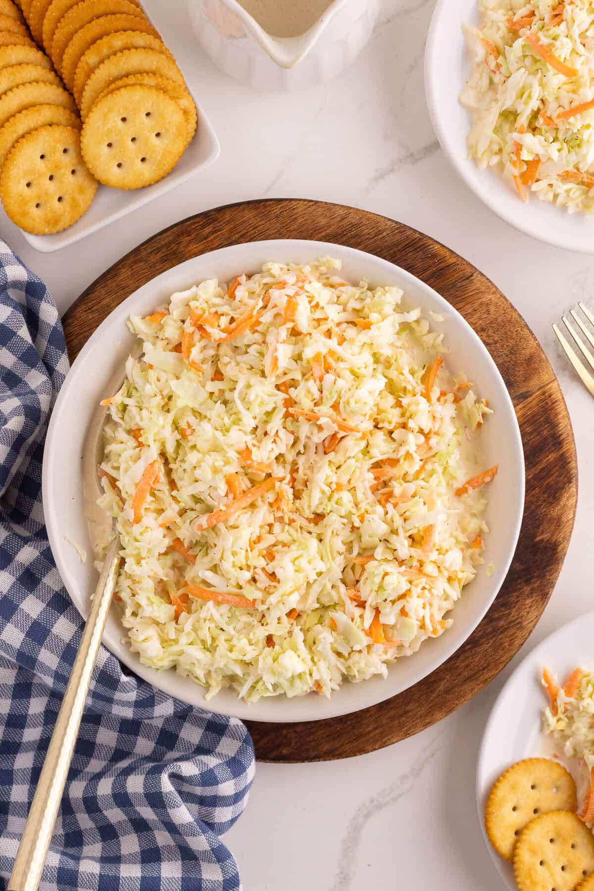 Classic coleslaw in a white serving bowl on a wood board with  small plates, crackers, and a blue gingham napkin.