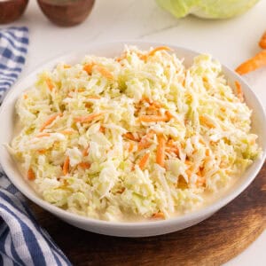 Creamy coleslaw in a white bowl on a wood board with a head of cabbage and carrots in the background.