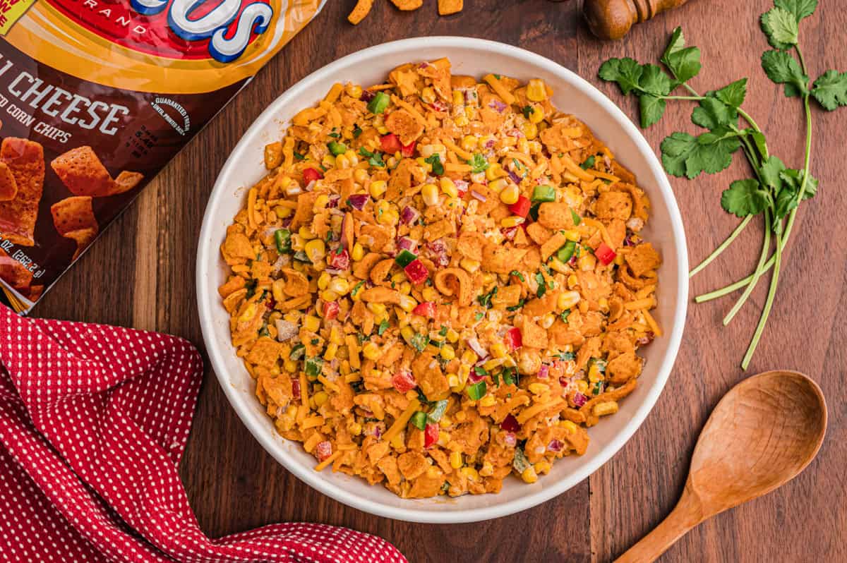 Overhead bowl of Frito salad next to a Chili Cheese Fritos bag, fresh cilantro, a wooden spoon, and a red gingham napkin on a wood board.