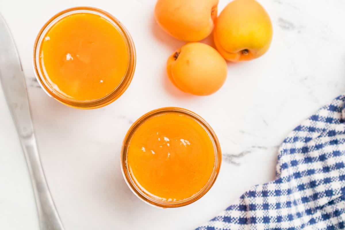 Two open jars of homemade apricot jam shot from above with fresh apricots, a knife, and a blue checked towel.