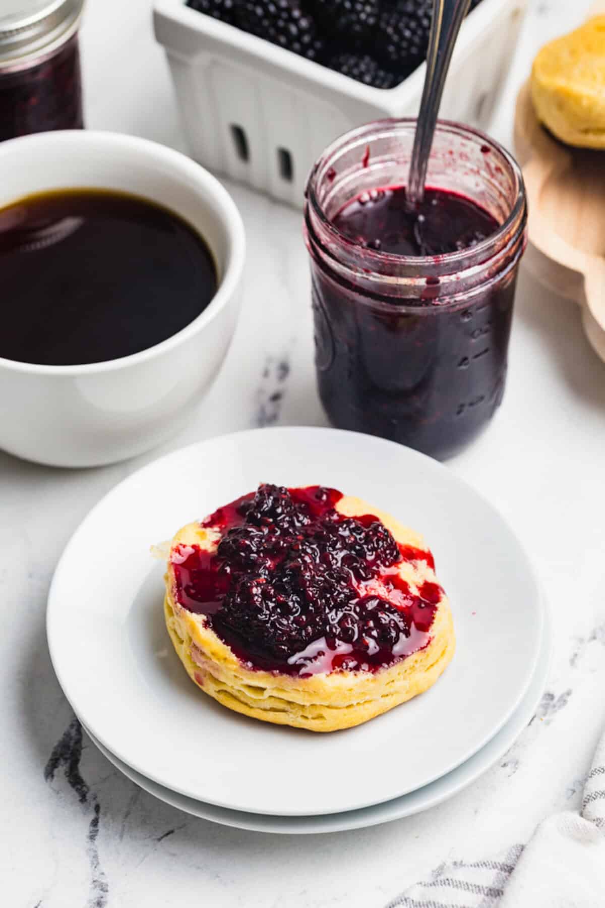 A split biscuit topped with blackberry jam on a white plate with a jar of jam and a cup of coffee in the background.