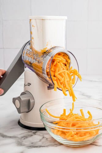 Hand crank rotary cheese shredder shredding cheddar into a bowl.
