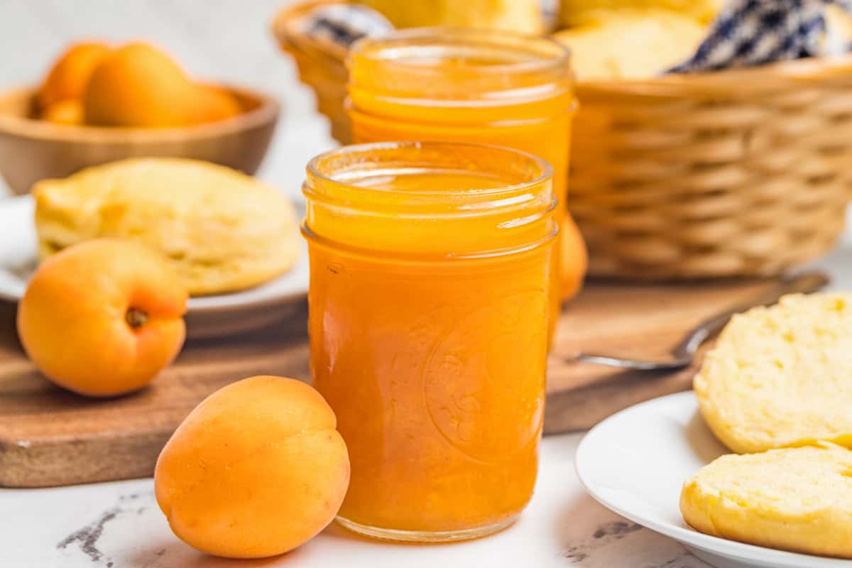 An open mason jar of homemade apricot jam on a wooden board with fresh apricots around it.