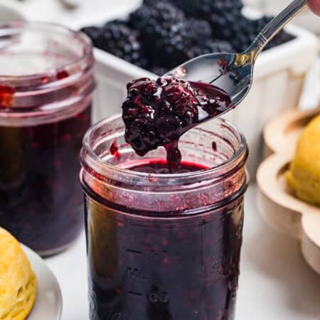 A spoon lifting a thick scoop of blackberry jam from a mason jar with a basket of fresh blackberries behind it.