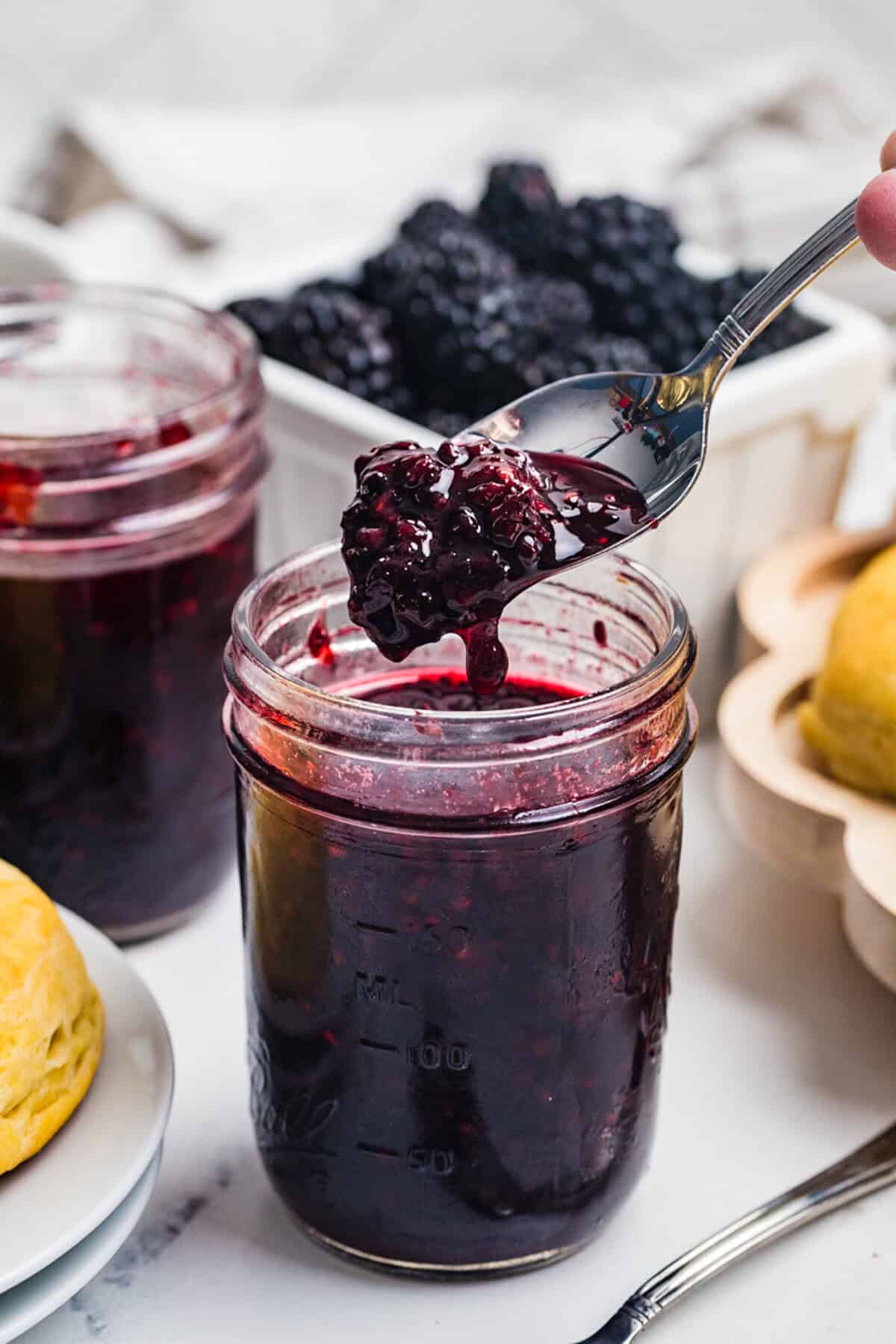 A spoon lifting a thick scoop of blackberry jam from a mason jar with a basket of fresh blackberries behind it.