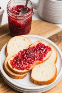 A slice of bread spread with raspberry jam on a white plate with a jar of homemade raspberry jam.