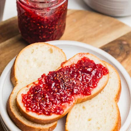 A slice of bread spread with raspberry jam on a white plate with a jar of homemade raspberry jam.