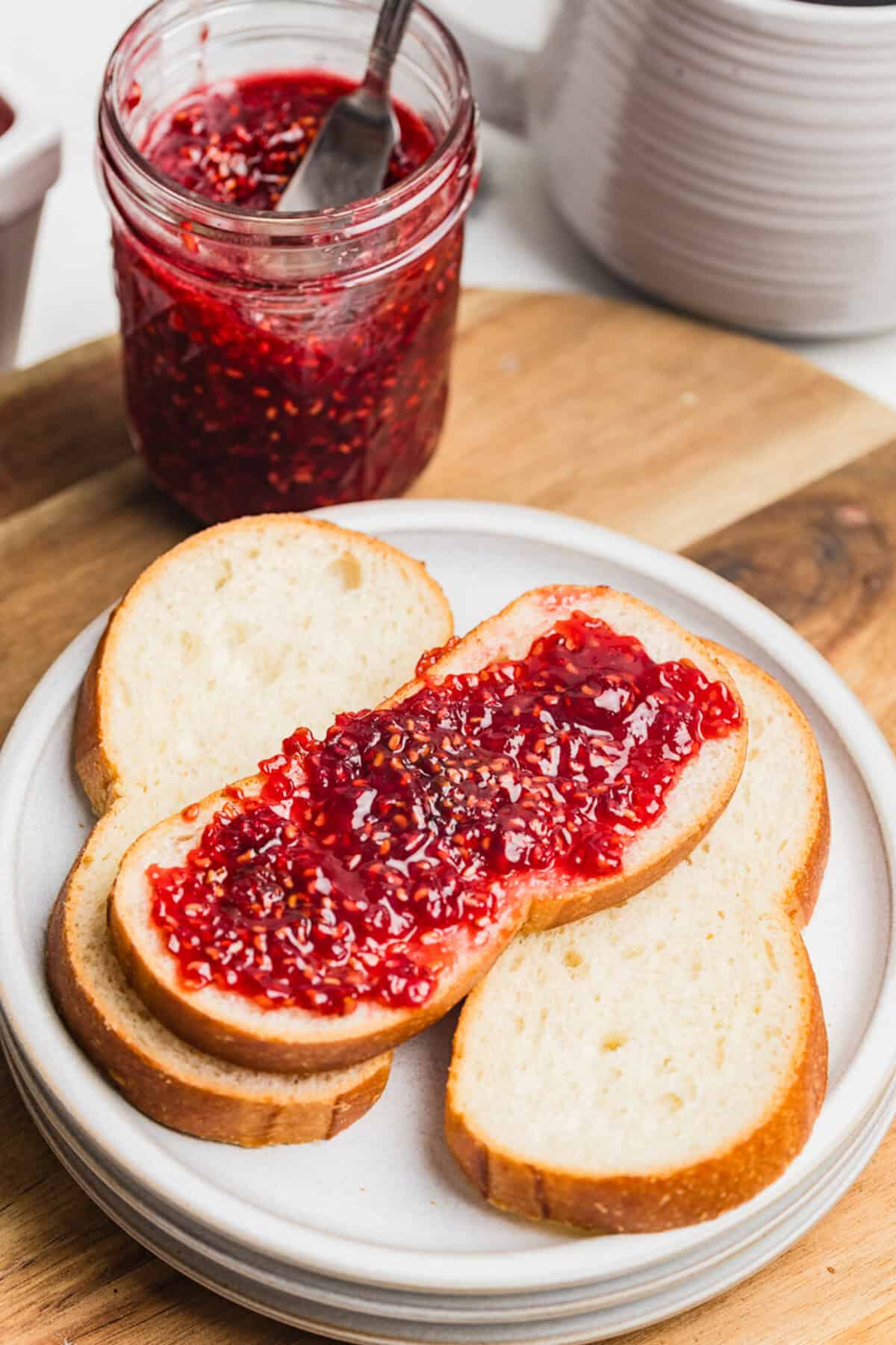 A slice of bread spread with raspberry jam on a white plate with a jar of homemade raspberry jam.
