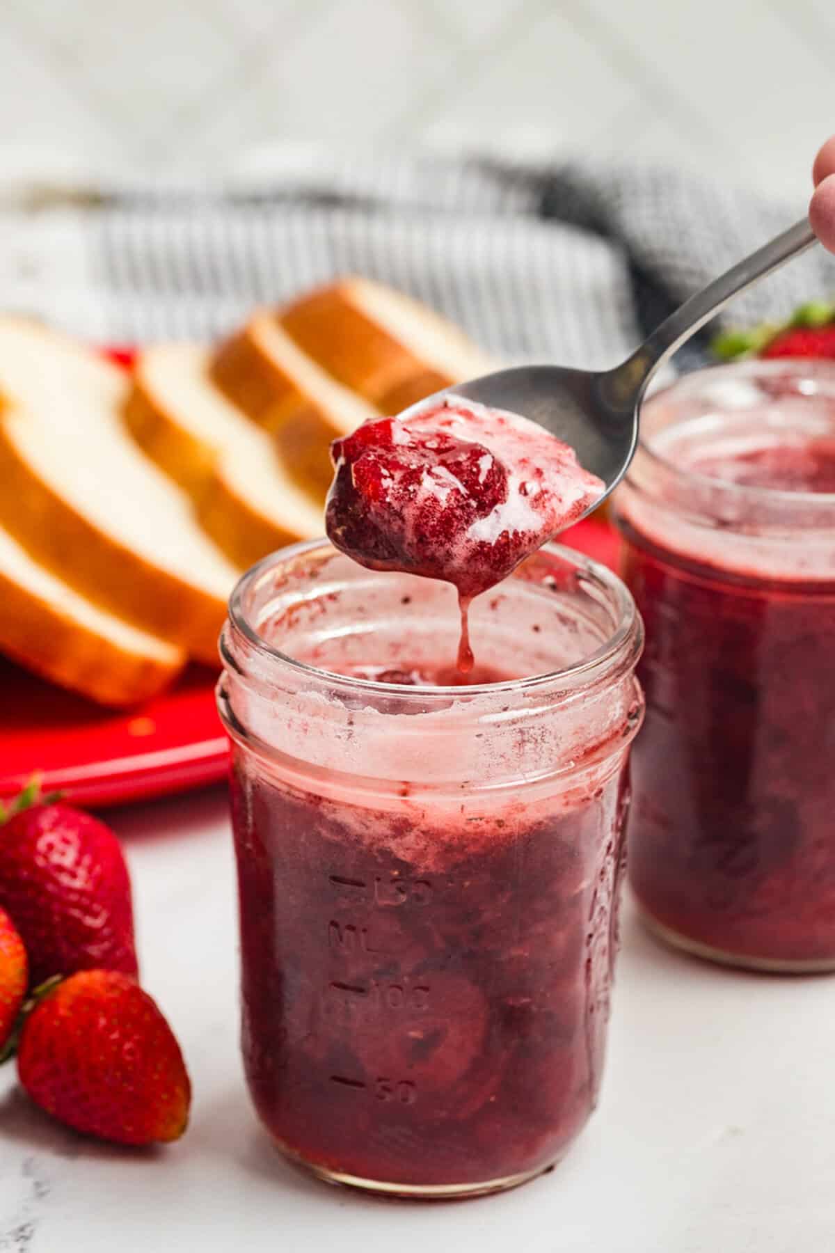 A spoon lifting strawberry jam from an open mason jar with bread slices nearby.