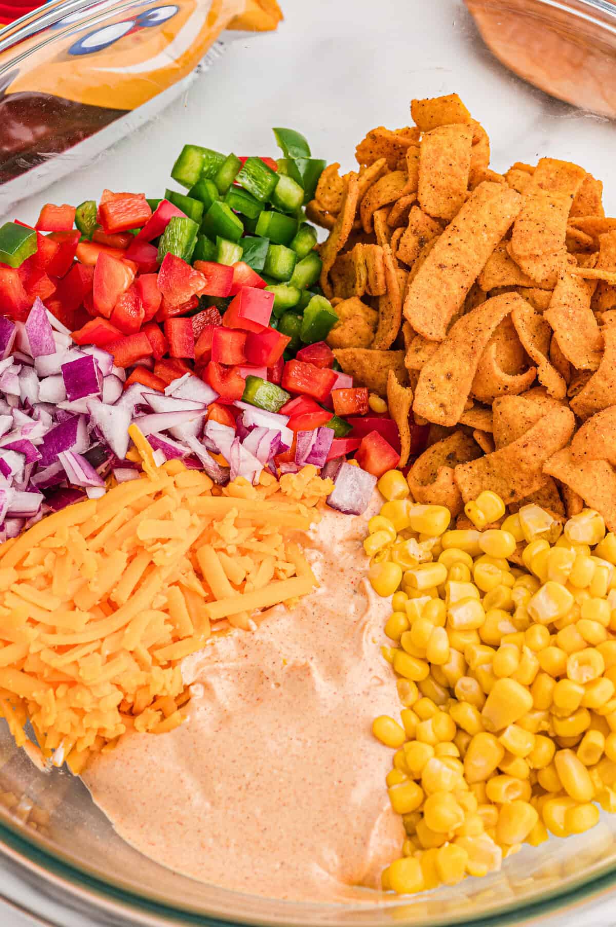 Glass bowl with diced bell pepper, red onion, shredded cheddar, sweet corn, Chili Cheese Fritos, and taco dressing arranged in sections before stirring.
