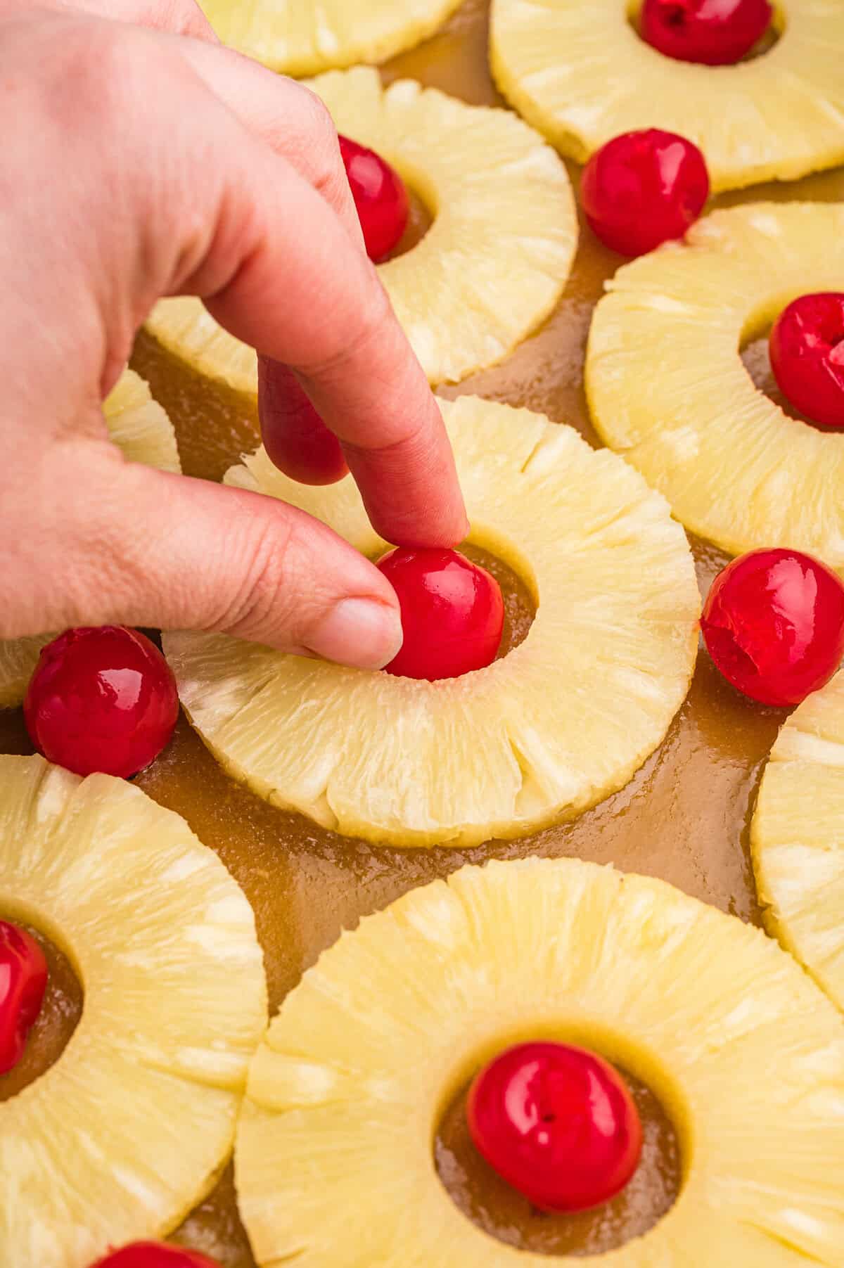 A hand placing a maraschino cherry into the center of a pineapple ring among other arranged rings in a pan.