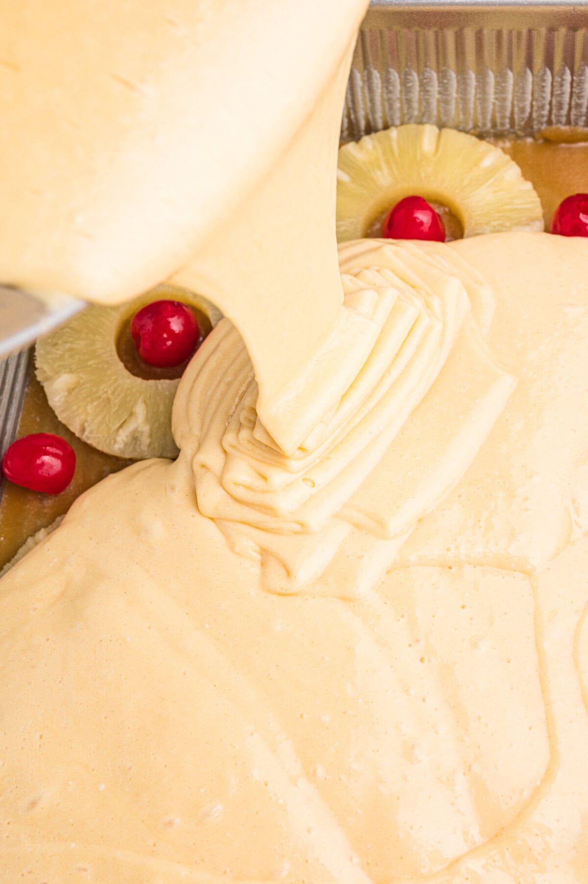 Yellow cake batter being poured over pineapple slices and cherries in a 9x13 baking pan.