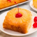 Square plated slice of pineapple upside down cake with a pineapple ring and a cherry on a white dish.