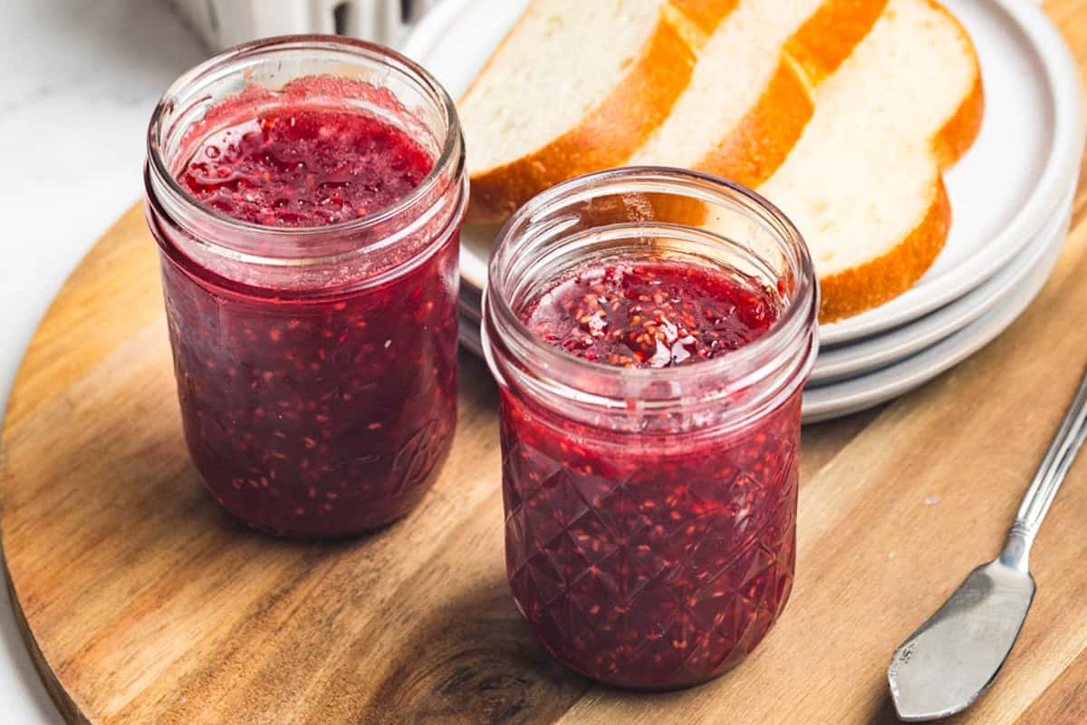 Two small mason jars filled with raspberry jam on a wooden board next to a stack of bread slices.