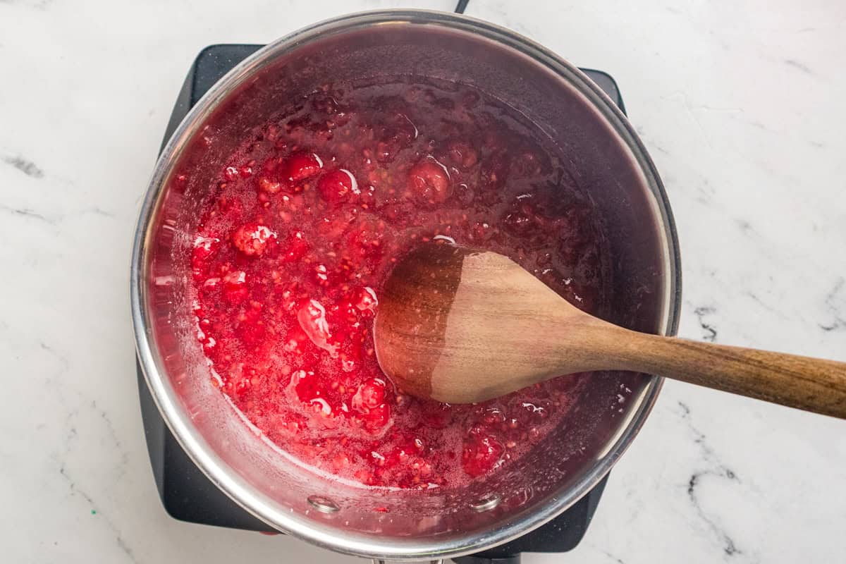 Raspberries breaking down in a saucepan with a wooden spoon stirring the bright red juice.