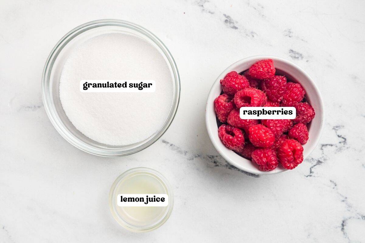 Ingredients for raspberry jam labeled on a marble counter: granulated sugar, fresh raspberries, and lemon juice.