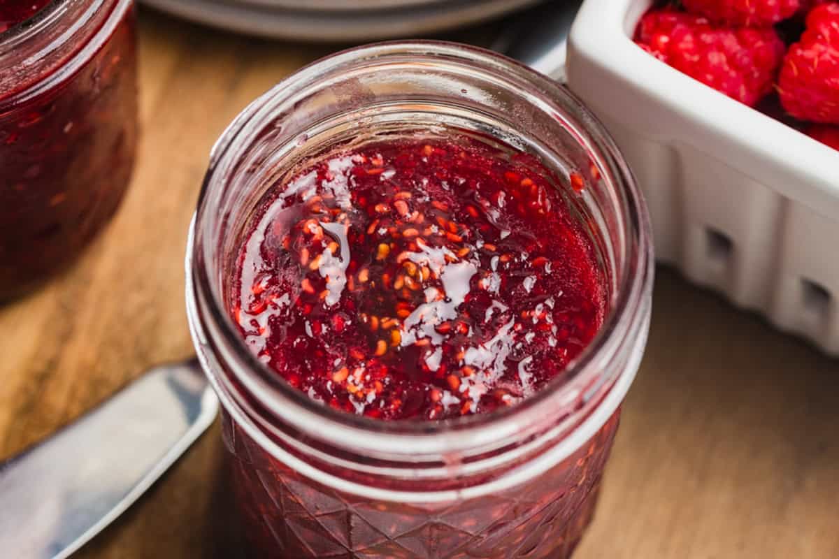 A slice of bread spread with raspberry jam on a white plate with a jar of jam and a basket of fresh raspberries to the side.
