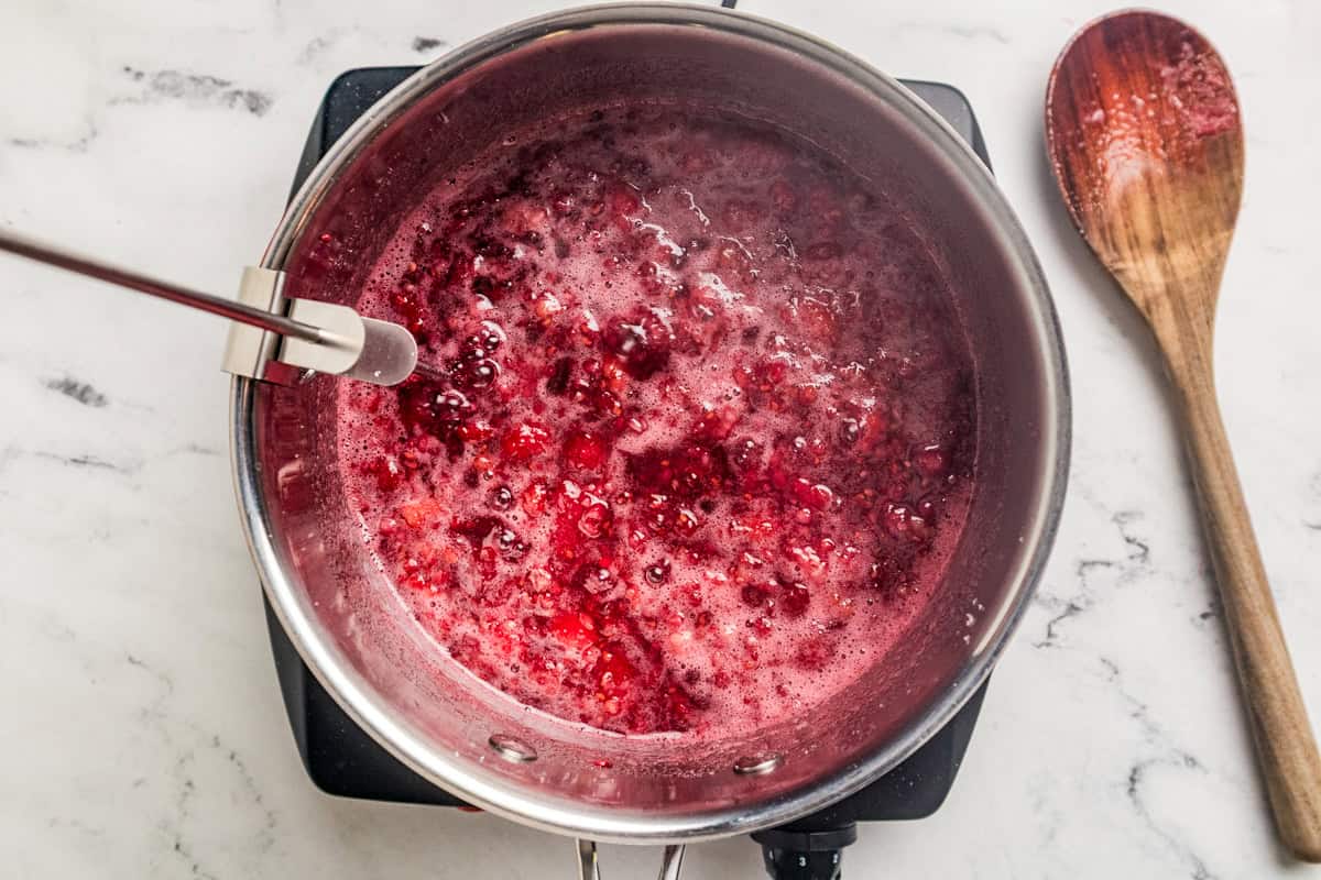 Bubbling raspberry jam in a saucepan with a candy thermometer attached and a wooden spoon to the side.