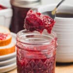 A close-up square shot of a spoon lifting raspberry jam from a small mason jar with a slice of bread in the background.