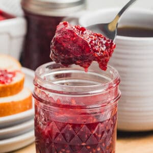 A close-up square shot of a spoon lifting raspberry jam from a small mason jar with a slice of bread in the background.
