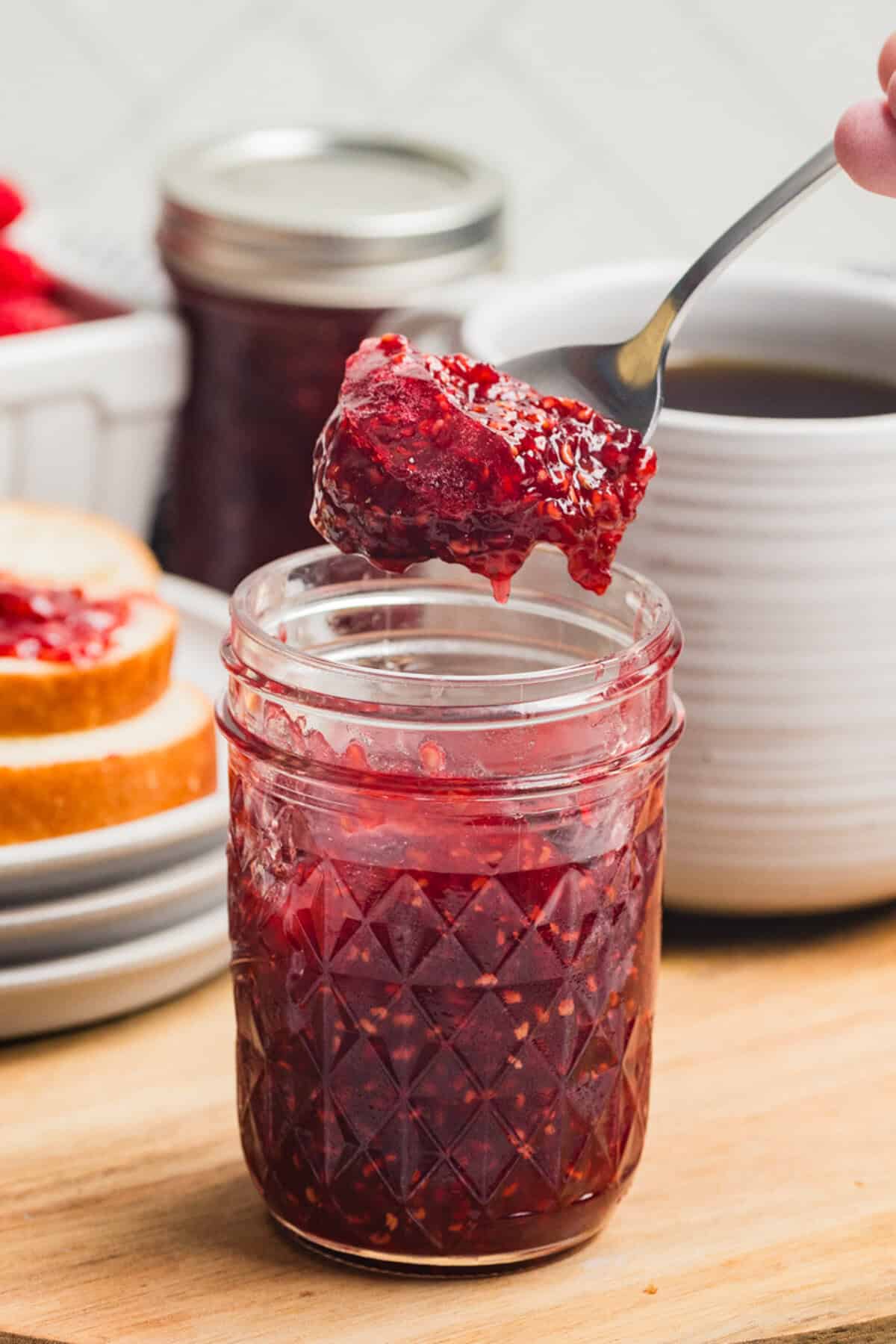 A spoon lifting raspberry jam from a small mason jar with a slice of bread and jar of jam in the background and a coffee mug to the side.