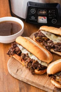 Three French dip sandwiches on a wooden cutting board with a bowl of au jus, with a Crock-Pot brand slow cooker visible in the background.