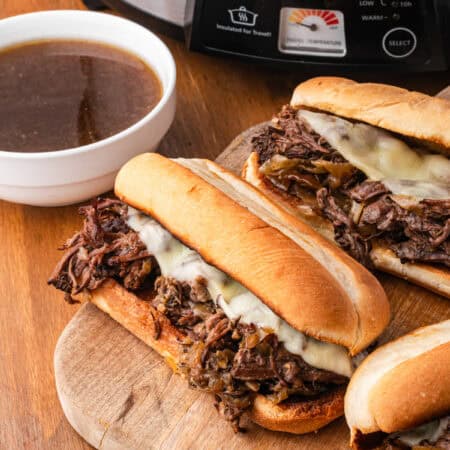 Three French dip sandwiches on a wooden cutting board with a bowl of au jus, with a Crock-Pot brand slow cooker visible in the background.