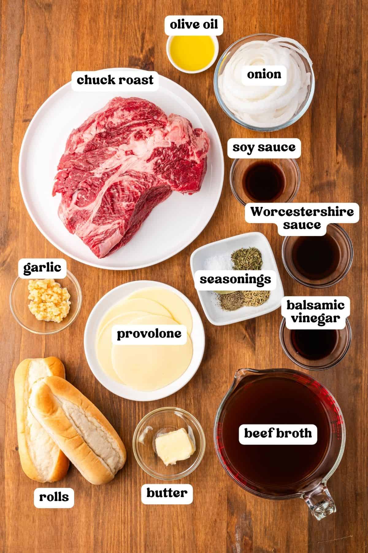 Labeled ingredients on a wood surface: chuck roast, olive oil, onion, garlic, beef broth, Worcestershire sauce, soy sauce, balsamic vinegar, seasonings, provolone, butter, and rolls.