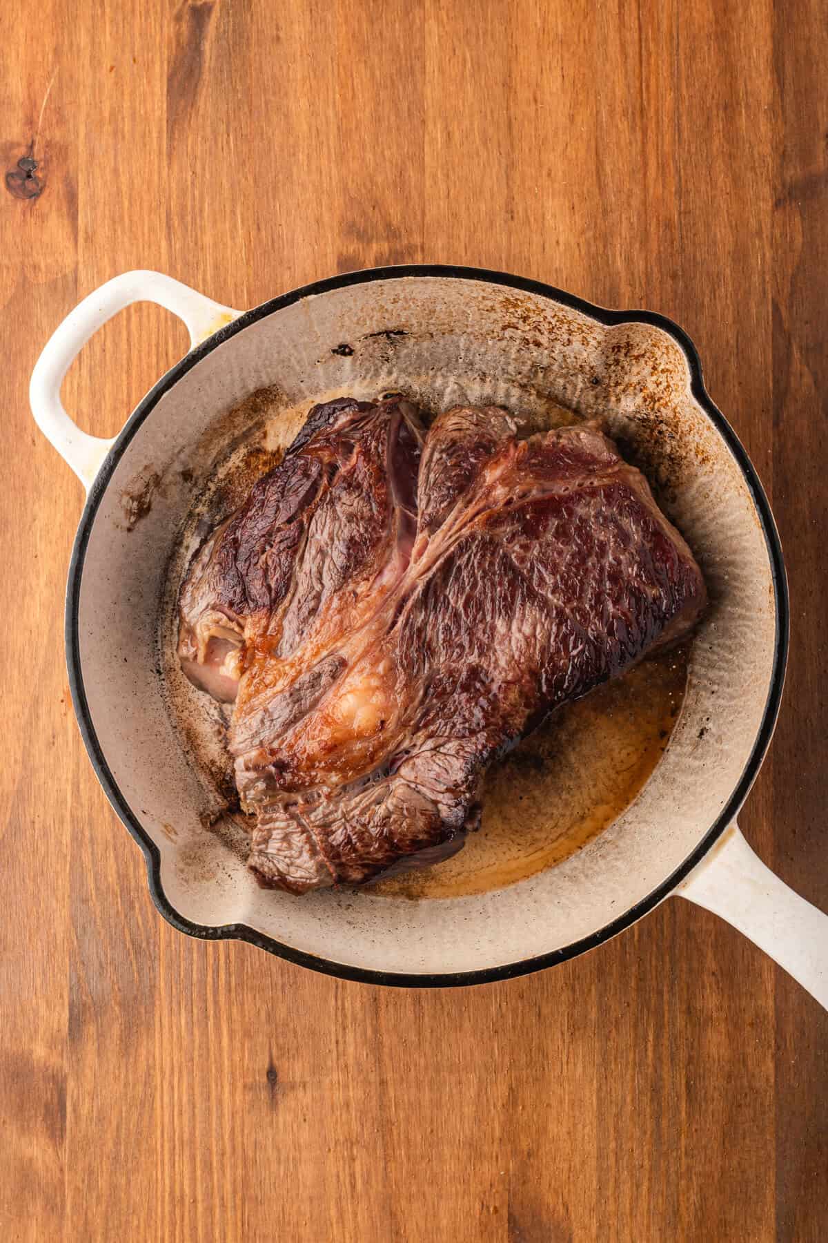 Top-down view of a seared chuck roast in a white Dutch oven on a wood surface.