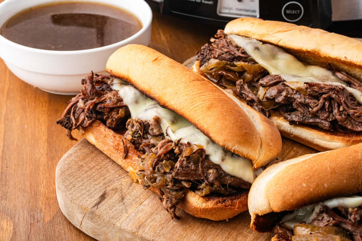Three French dip sandwiches on a wooden cutting board with a white bowl of au jus on the side and a slow cooker partially visible in the background.
