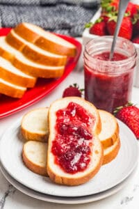 A slice of bread topped with strawberry jam in front of a red platter of bread slices, a jar of jam with a spoon, and fresh strawberries.