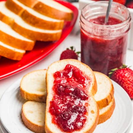A slice of bread topped with strawberry jam in front of a red platter of bread slices, a jar of jam with a spoon, and fresh strawberries.