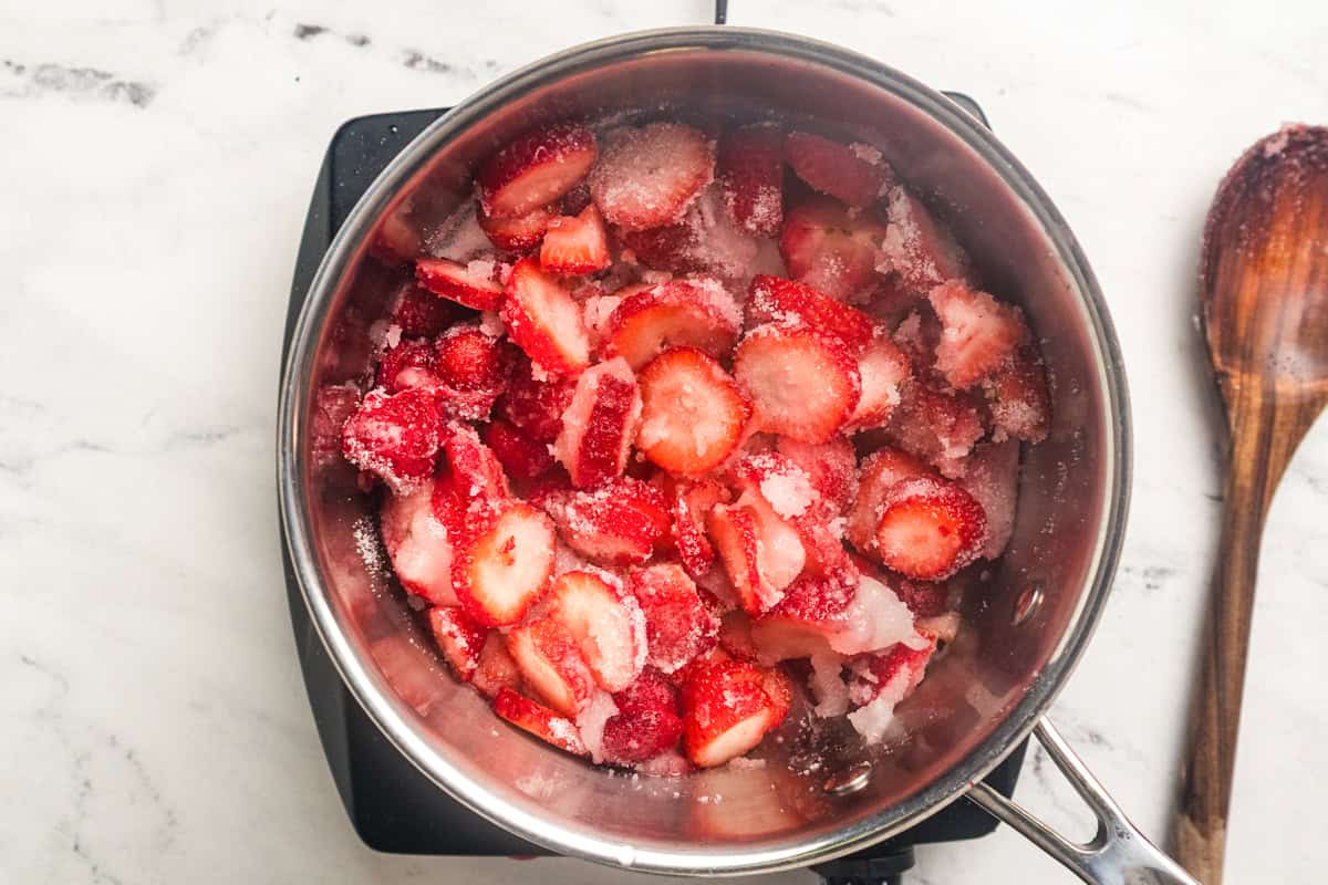 Sliced strawberries in a saucepan sprinkled with granulated sugar with a wooden spoon to the side.