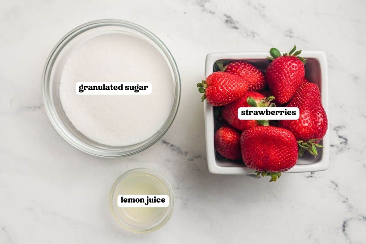 Ingredients for strawberry jam labeled on a marble counter: granulated sugar, fresh strawberries, and lemon juice.