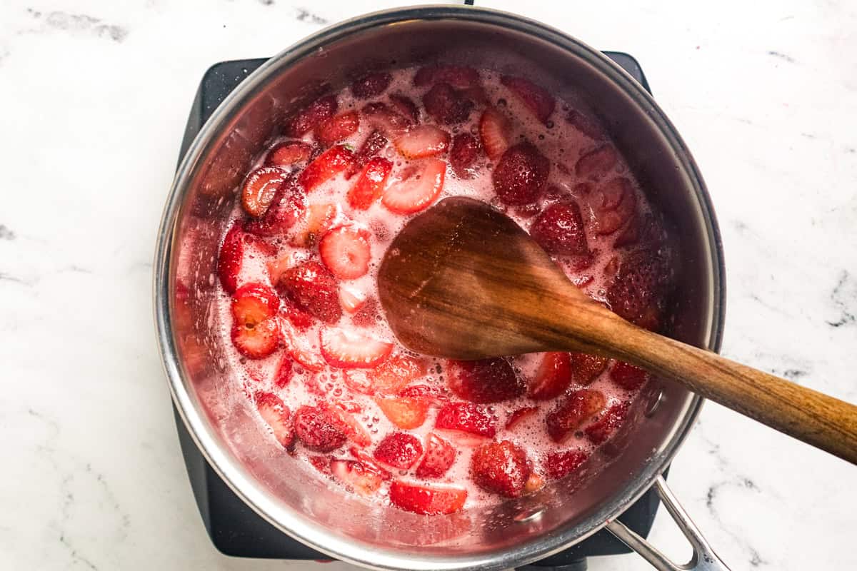Softened sliced strawberries simmering in a saucepan with a wooden spoon stirring the bright pink mixture.