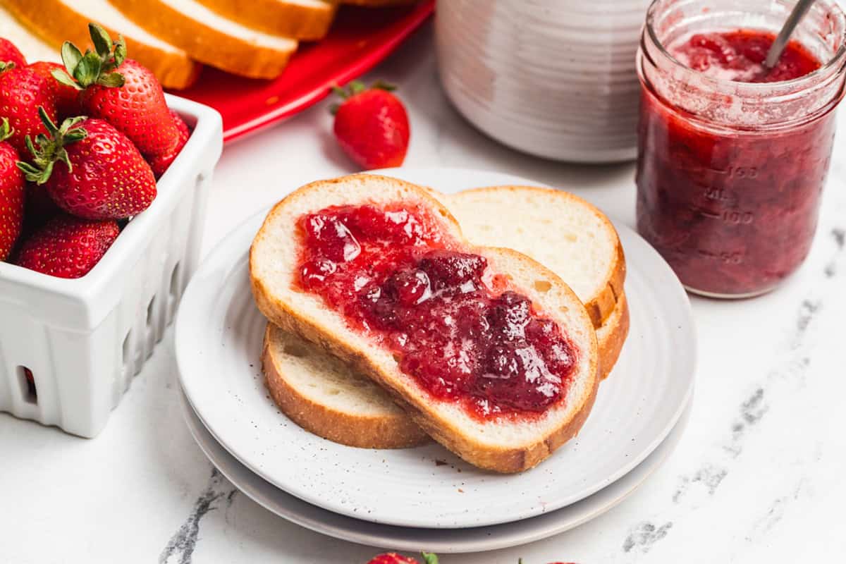 A bread slice topped with strawberry jam on a red strawberry-shaped plate next to a jar of jam and fresh strawberries.
