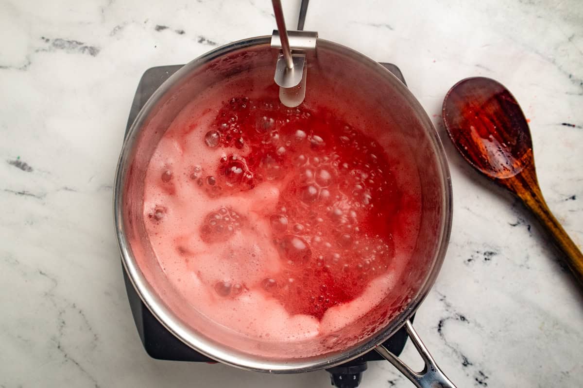 Bubbling strawberry jam in a saucepan with a candy thermometer attached to the rim.