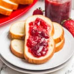 Bread slices topped with homemade strawberry jam stacked on a white plate with a jar of jam nearby.