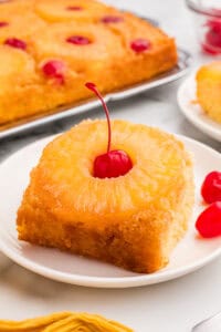 A square slice of pineapple upside down cake on a white plate with a pineapple ring and a maraschino cherry on top.