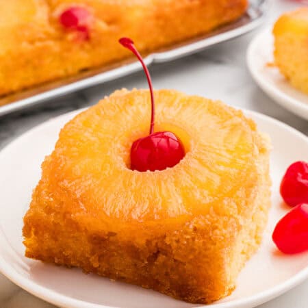 A square slice of pineapple upside down cake on a white plate with a pineapple ring and a maraschino cherry on top.