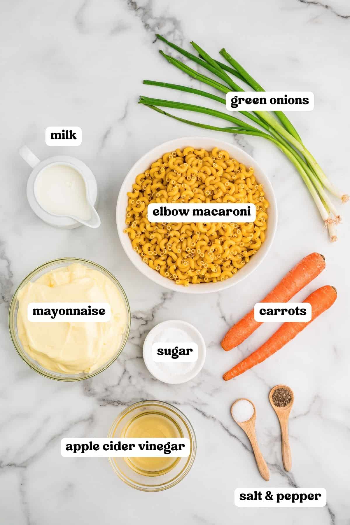 Labeled ingredients on a marble counter: green onions, milk, elbow macaroni, mayonnaise, sugar, carrots, apple cider vinegar, and salt and pepper.