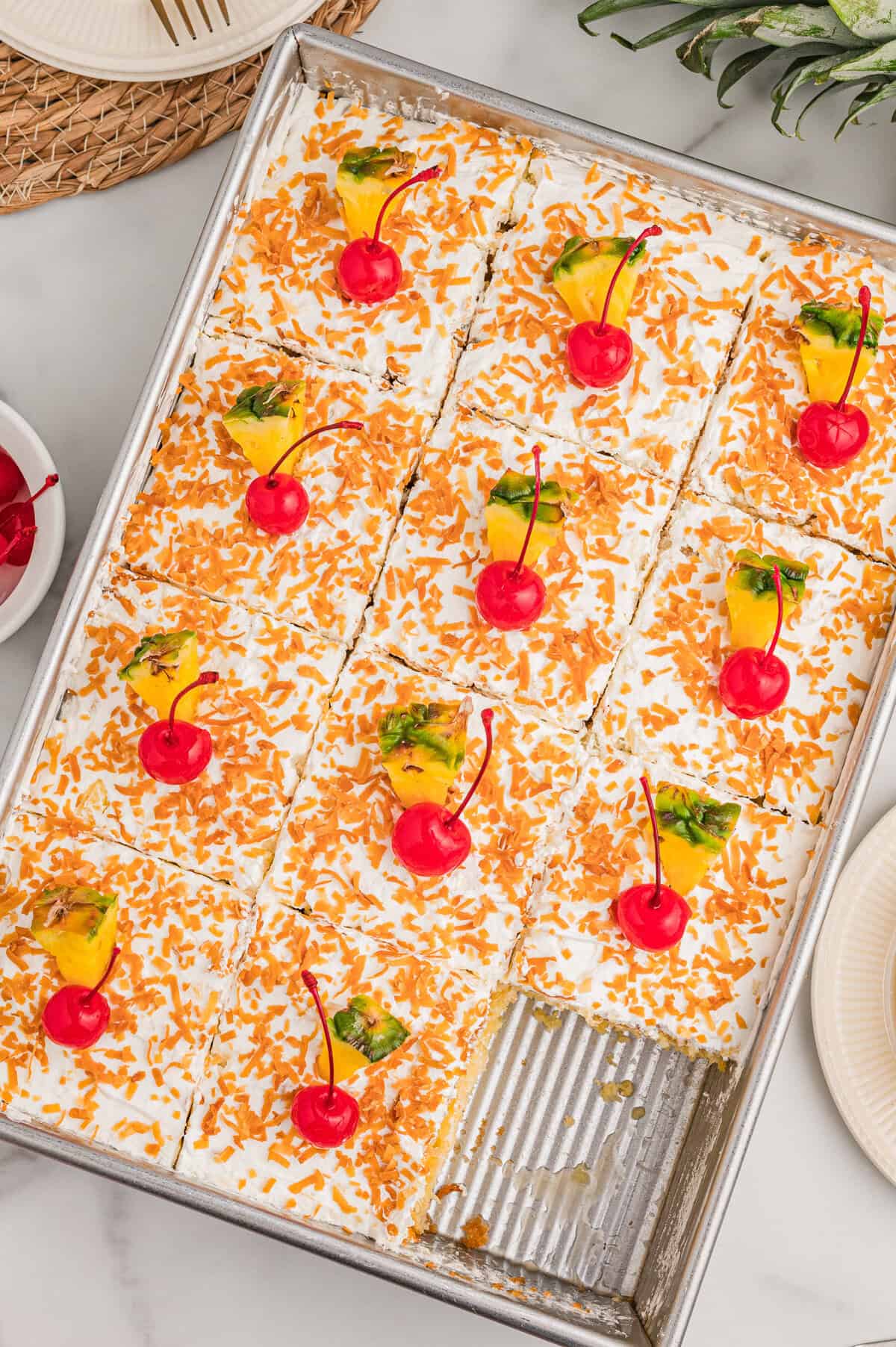 Top-down view of the cake in a pan with one corner piece removed, squares topped with toasted coconut, cherries, and pineapple.