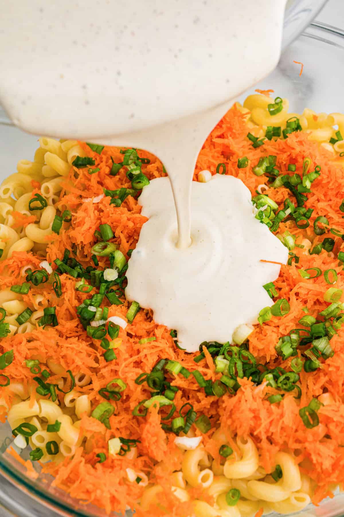 Creamy dressing being poured over cooked macaroni, shredded carrots, and green onions in a large glass bowl.
