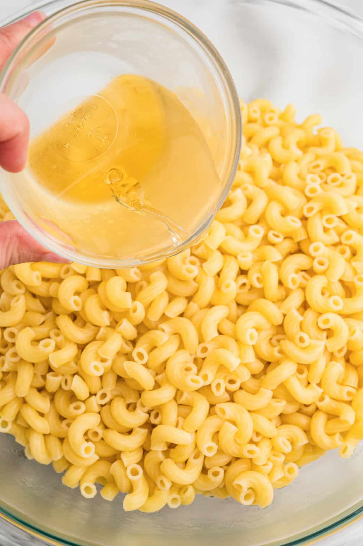 Apple cider vinegar being poured from a small glass bowl over cooked elbow macaroni in a large glass mixing bowl.