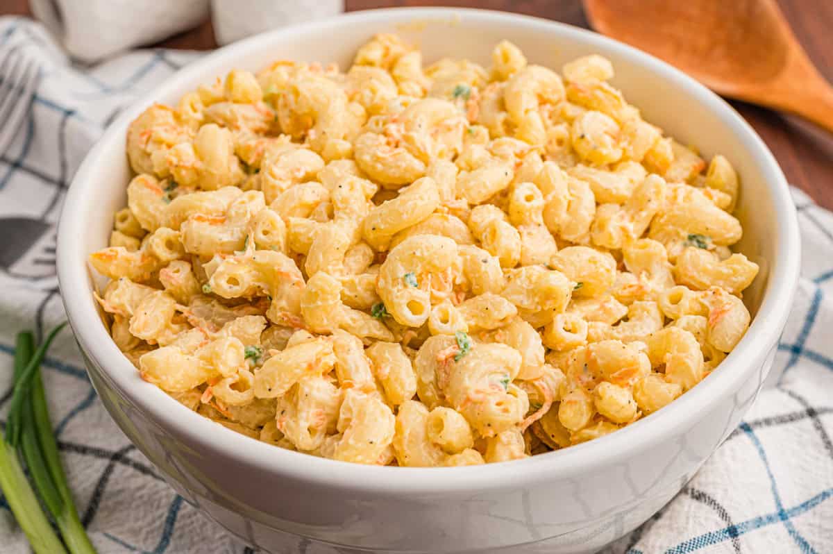 A white bowl of Hawaiian mac salad on a checkered dish towel with green onions and a wooden spoon nearby.