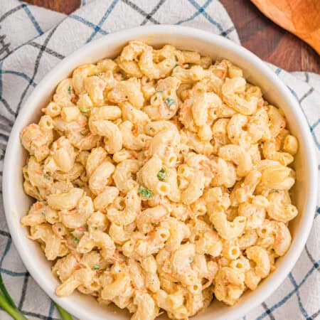 Top-down view of a white bowl of creamy macaroni salad on a checkered dish towel with green onions and a wooden spoon on the side.