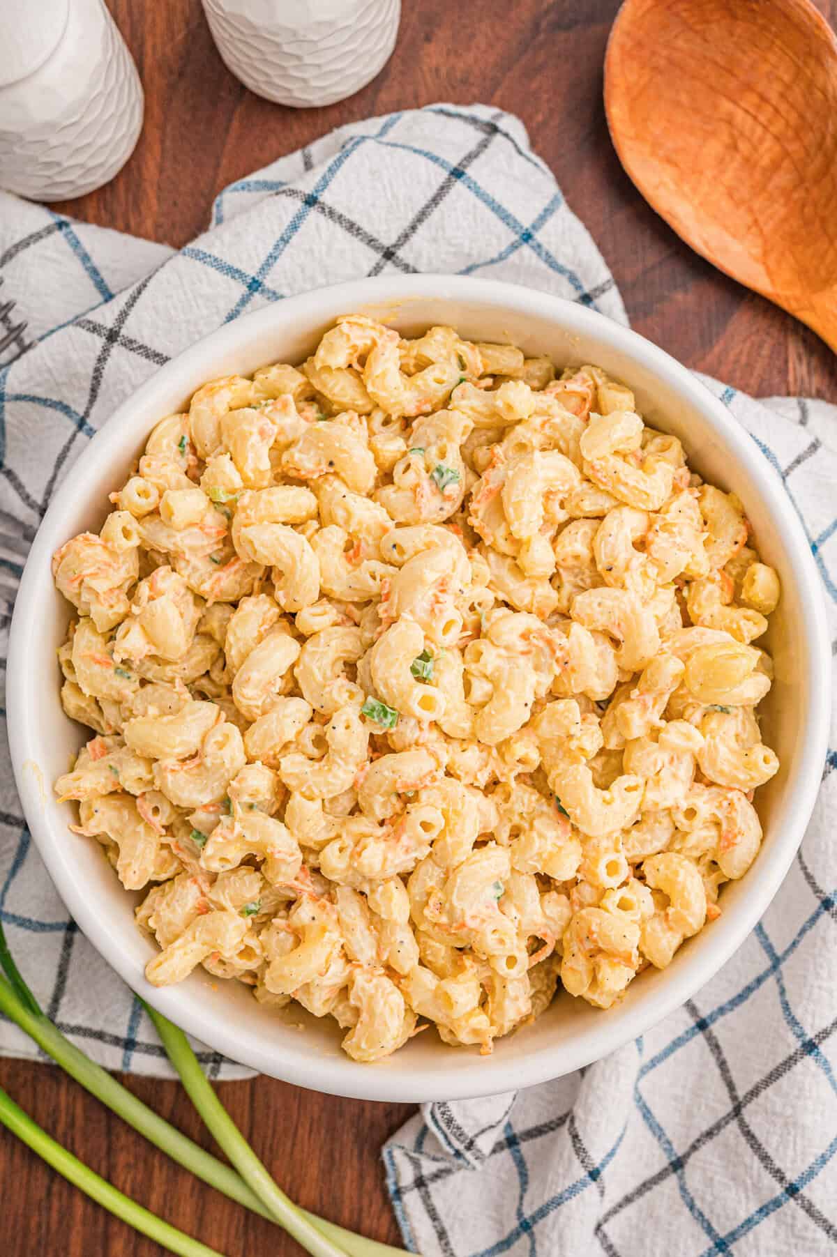 Top-down view of a white bowl of creamy macaroni salad on a checkered dish towel with green onions and a wooden spoon on the side.
